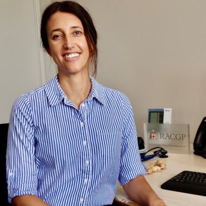 Dr Catherine Bourke smiles at the camera whilst sitting at her desk. She has dark hair which is in a ponytail and is wearing a white and blue striped shirt