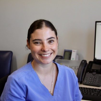 Dr Chelsea May smiles at the camera. She had dark hair which is pulled back into a ponytail. She is sat at her desk and is wearing a powder blue scrub top.