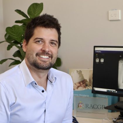 Dr Chris Hall smiles at the camera. He has short dark hair and dark facial hair. He is sat at his desk and is wearing a light blue shirt