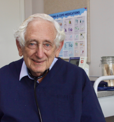Dr Colin Binns smiles at the camera. He is sitting in a clinical setting and has short white hair. He is wearing a pale blue long sleeve shirt with a darker blue short sleeve top on top.