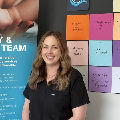 Dr Imogen Price smiles at the camera. She has long brown wavy hair and is wearing a black scrub top. She is standing in front of a Quinns Mindarie Super Clinic banner.