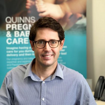 Dr Joshua Ebsworthy smiles at the camera. He has short dark hair and is wearing dark glasses. He is sat at his desk and is wearing a blue and white shirt