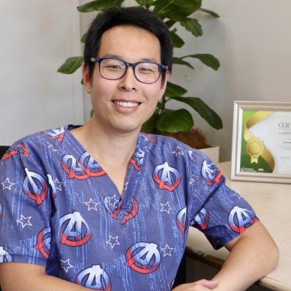 Dr Jonathan Chia smiles at the camera. He has short dark hair and is wearing dark round glasses. He is wearing Avengers scrubs and is sat at his desk with a certificate in sight.