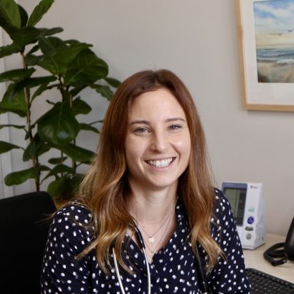 Dr Krischelle McCallum smiles at the camera. She had long reddish brown hair and is sat at her desk. She is wearing a black and white polka dot top with a stethoscope around her neck.