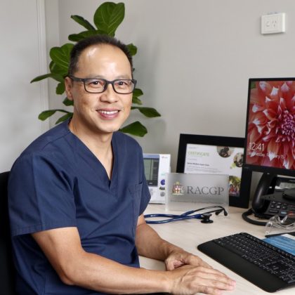 Dr Lawrence Chin smiles at the camera whilst sitting at his desk. He has short dark hair and is wearing grey glasses.