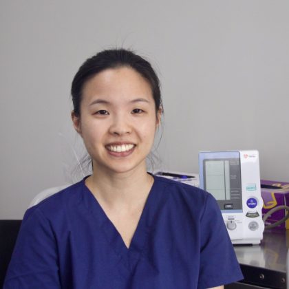 Dr Lisa Chau smiles at the camera. She is in a clinical setting and has black hair which is tied back. She is wearing a blue scrub top.