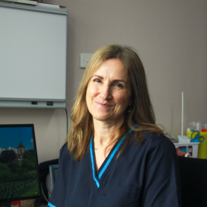 Dr Lyn Wren smiles at the camera. She has long brown hair and is in a clinical setting. She has a dark blue scrub top on.