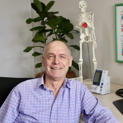 Dr Ross Dunstan smiles at the camera. He is in a clinical setting and is wearing a pink and blue striped shirt. He has short dark and grey hair.