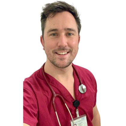 Dr Greg Bailey smiles at the camera. He has short brown hair and is wearing red scrubs with a stethescope around his neck