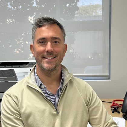 Dr Greg Bailey smiles at the camera. He has short dark hair and light facial hair. He is sat at his desk and is wearing a beige sweater