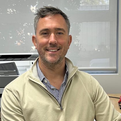 Dr Greg Bailey smiles at the camera. He has short dark hair and light facial hair. He is sat at his desk and is wearing a beige sweater